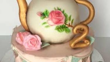 Beautiful tea kettle white with pink rose and gold trim on a light pink cake with 2 pink roses beside the kettle