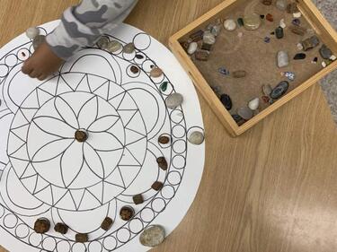 Early Learning, a child's hand placing rocks on a puzzle picture on a wooden desk