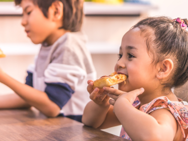 a boy and girl sitting on a wooden table next to each other eating pizza