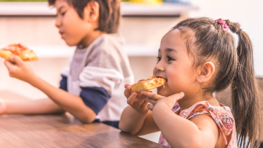 a boy and girl sitting on a wooden table next to each other eating pizza