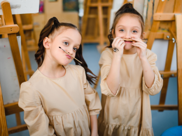 Twin girls dressed in a khaki coloured long sleeve dress with paint easles behind them holding paint brushes as mustaches on their faces