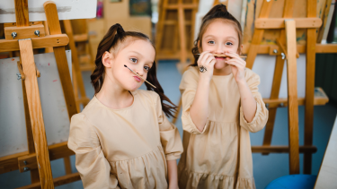 Twin girls dressed in a khaki coloured long sleeve dress with paint easles behind them holding paint brushes as mustaches on their faces