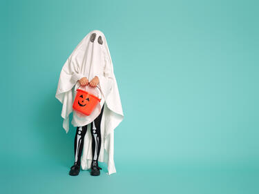 Child wearing ghost costume posing against turquoise background holding a pumpkin candy container
