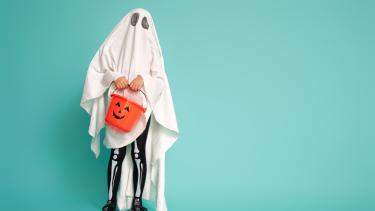 Child dressed up as a ghost for halloween Child wearing ghost costume posing against turquoise background holding a pumpkin candy container