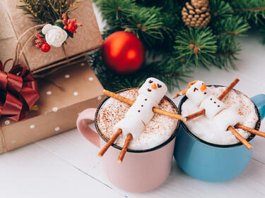 a pink and blue mug with hot chocolate and snowman treats on top next to presents and christmas tree
