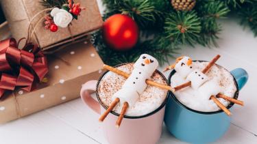 a pink and blue mug with hot chocolate and snowman treats on top next to presents and christmas tree